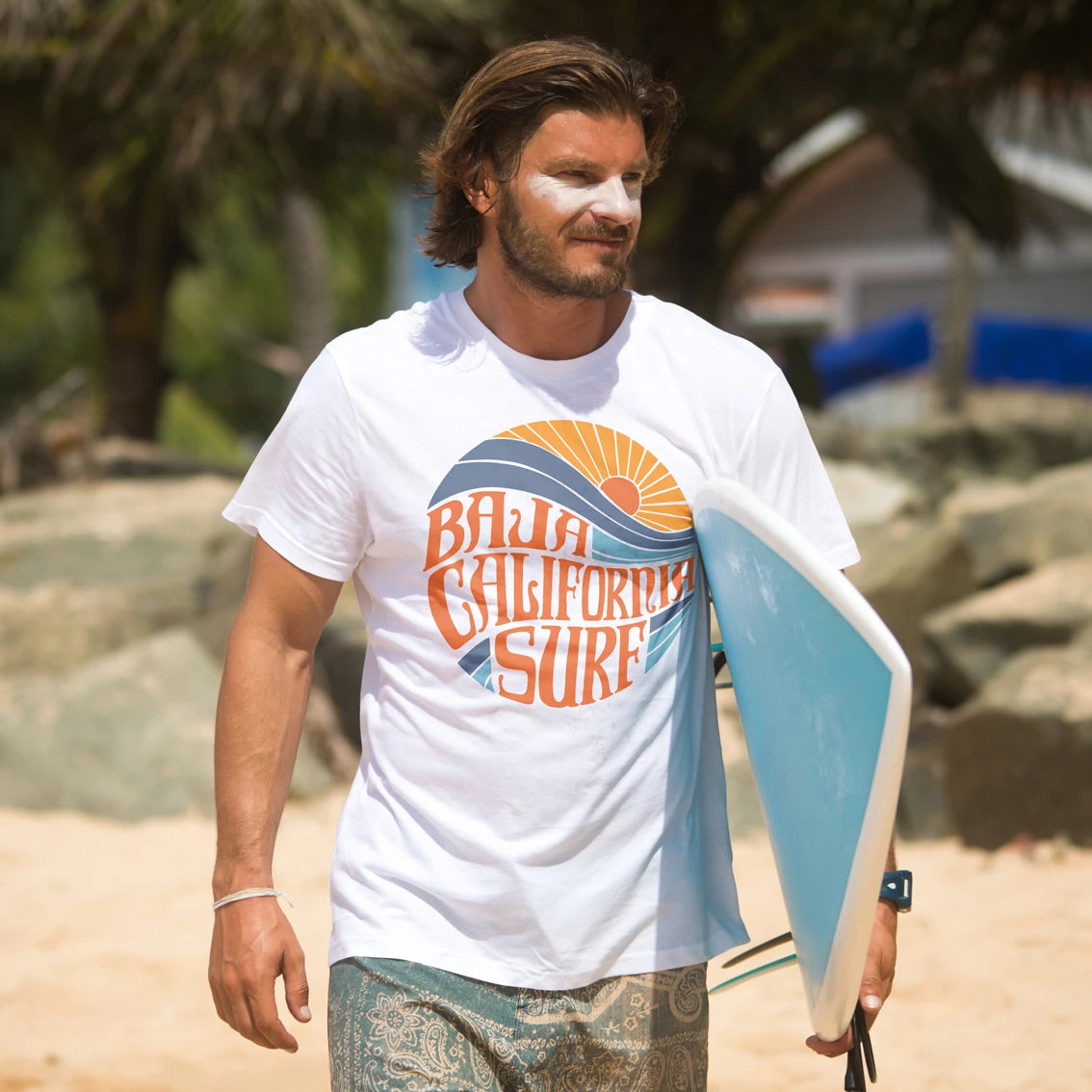 A man walking on a sandy beach carrying a light blue surfboard, wearing a white graphic t-shirt that reads 'Baja California Surf' in orange and blue, with palm trees and rocks in the background.