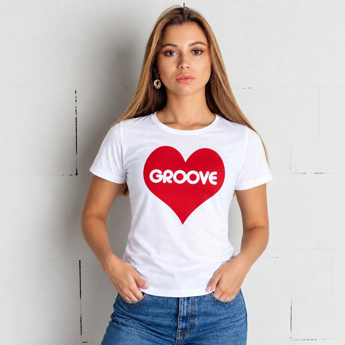 A woman wearing a white short-sleeve t-shirt with a red heart graphic on the chest containing the word 'GROOVE' in white, paired with blue jeans, standing against a white brick wall.