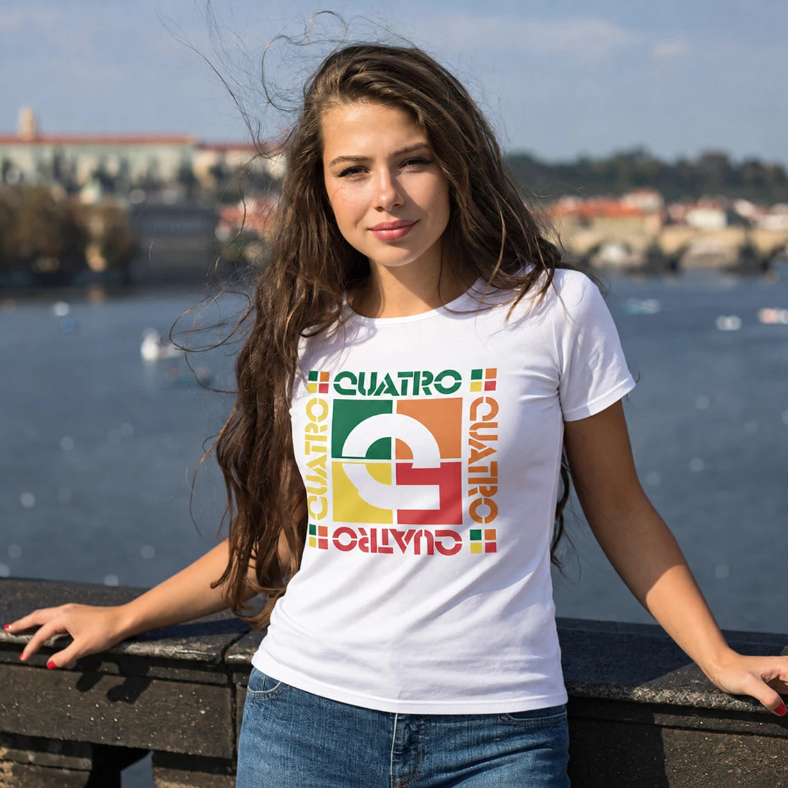 A woman wearing a white graphic t-shirt with a colorful retro design that reads 'QUATRO' in green, yellow, orange, and red, standing outdoors by a body of water with a blurred cityscape background.