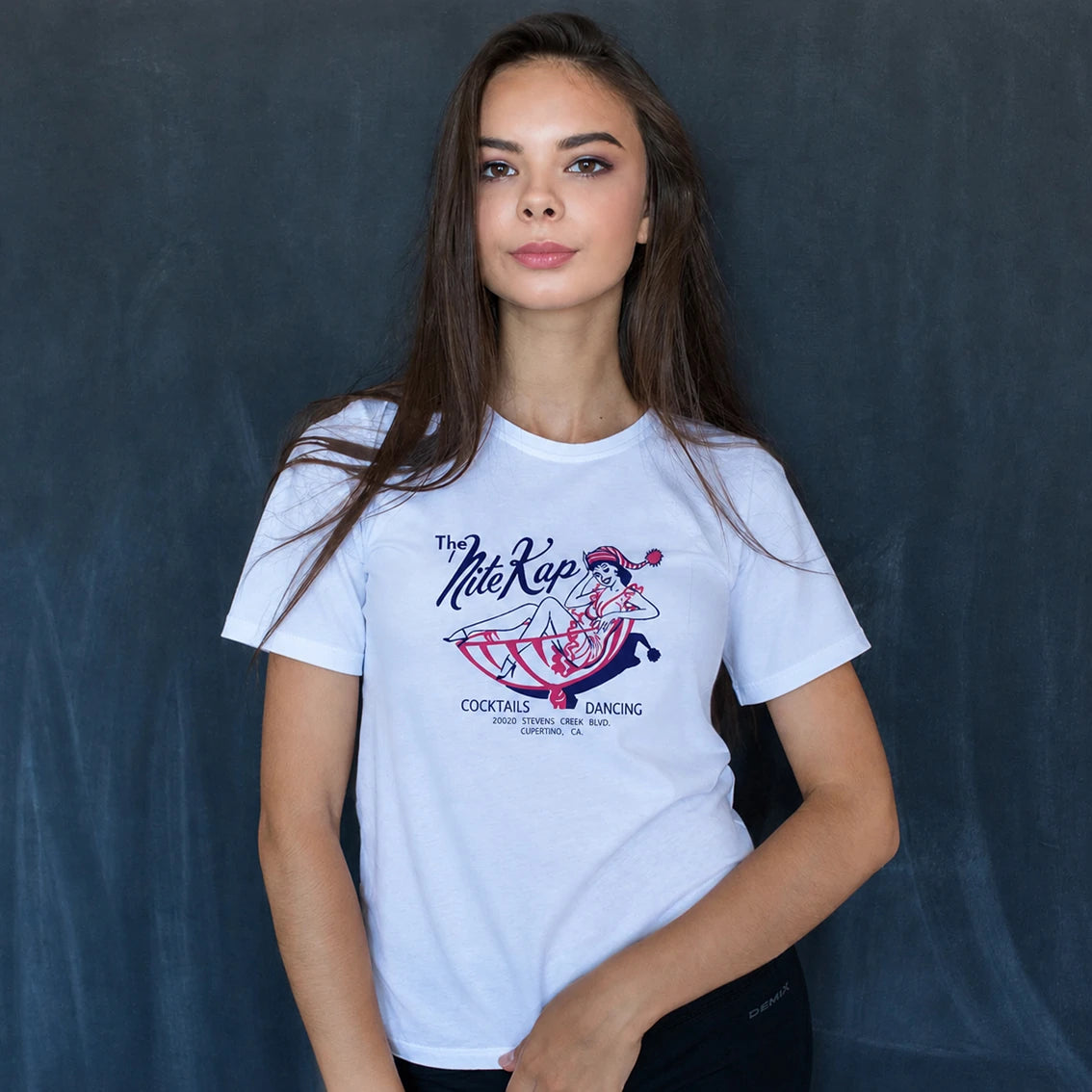 A woman modeling a white graphic t-shirt with a vintage-style 'The Nite Kap' cocktail graphic on the front, set against a dark textured studio backdrop.
