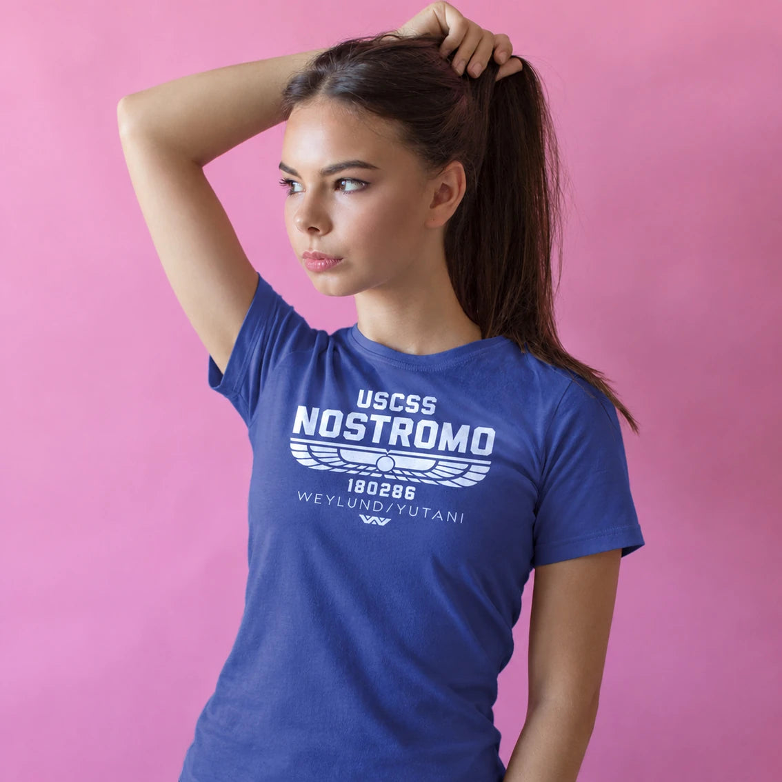 A woman wearing a royal blue crewneck t-shirt with a white graphic print that reads USCSS NOSTROMO standing against a solid pink background.