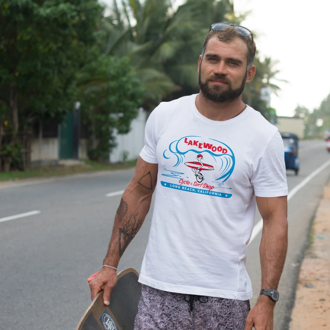 Man wearing a white t-shirt with a graphic design and 'Lakewood Cycle & Surf Shop' text, holding a skateboard on a road.