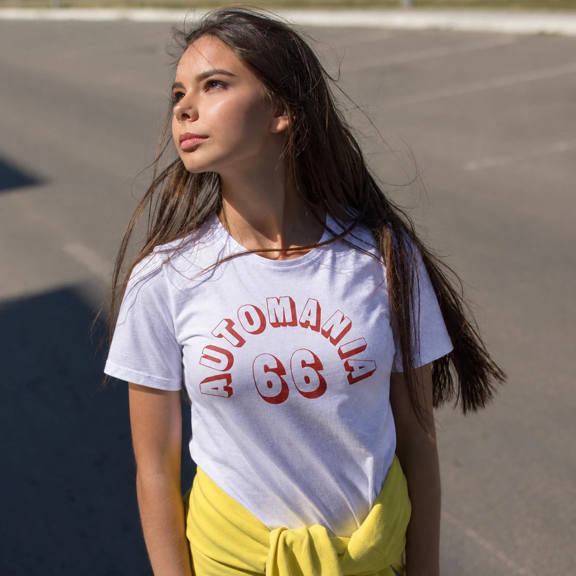 A woman wearing a white graphic t-shirt printed with 'AUTOMANIA 66', standing outdoors on a paved surface.