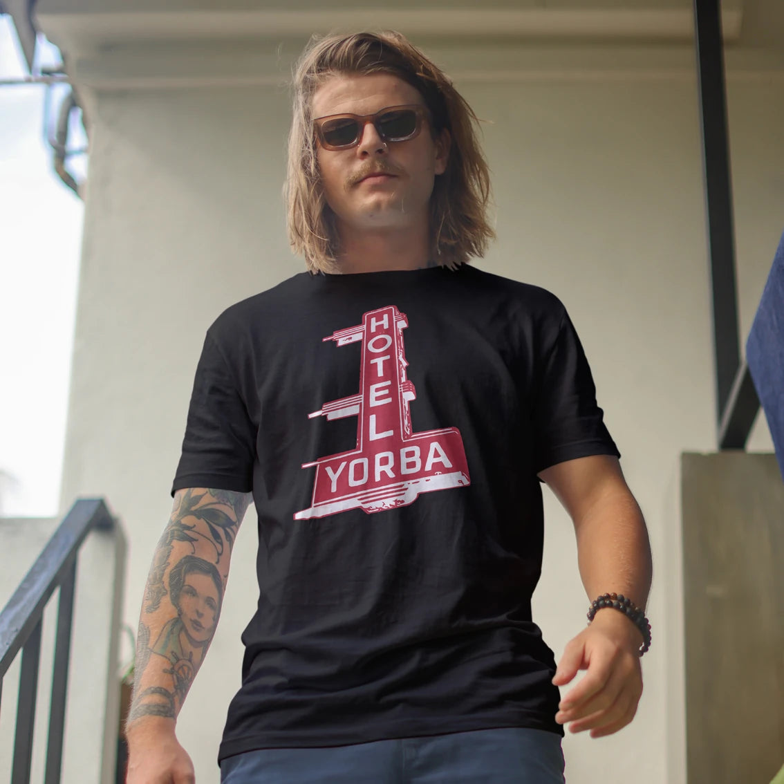 A man wearing a black t-shirt with a red and white retro sign graphic that reads 'HOTEL YORBA', standing outdoors against a light-colored building wall.
