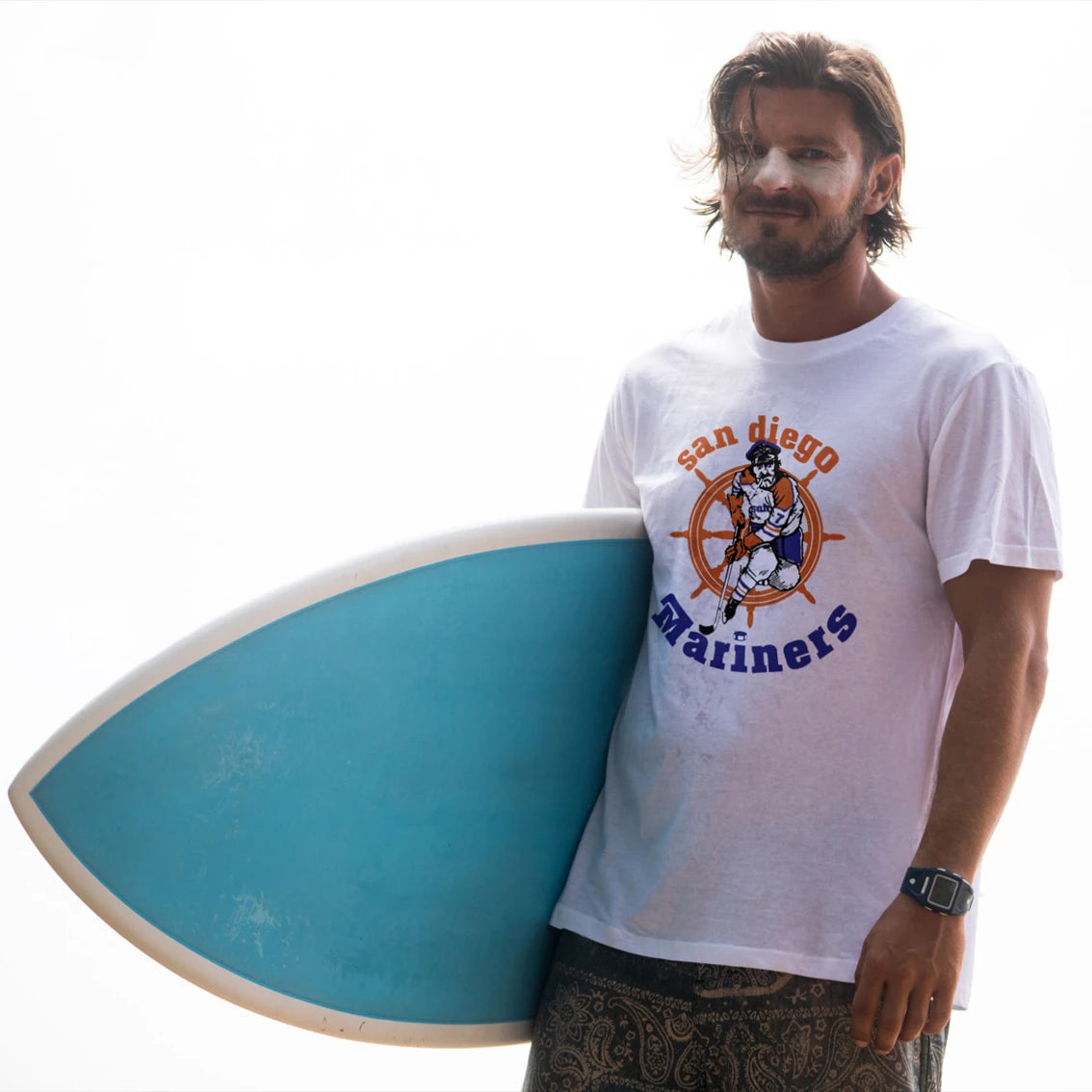 Man holding a blue surfboard wearing a 'San Diego Mariners' t-shirt on a white background