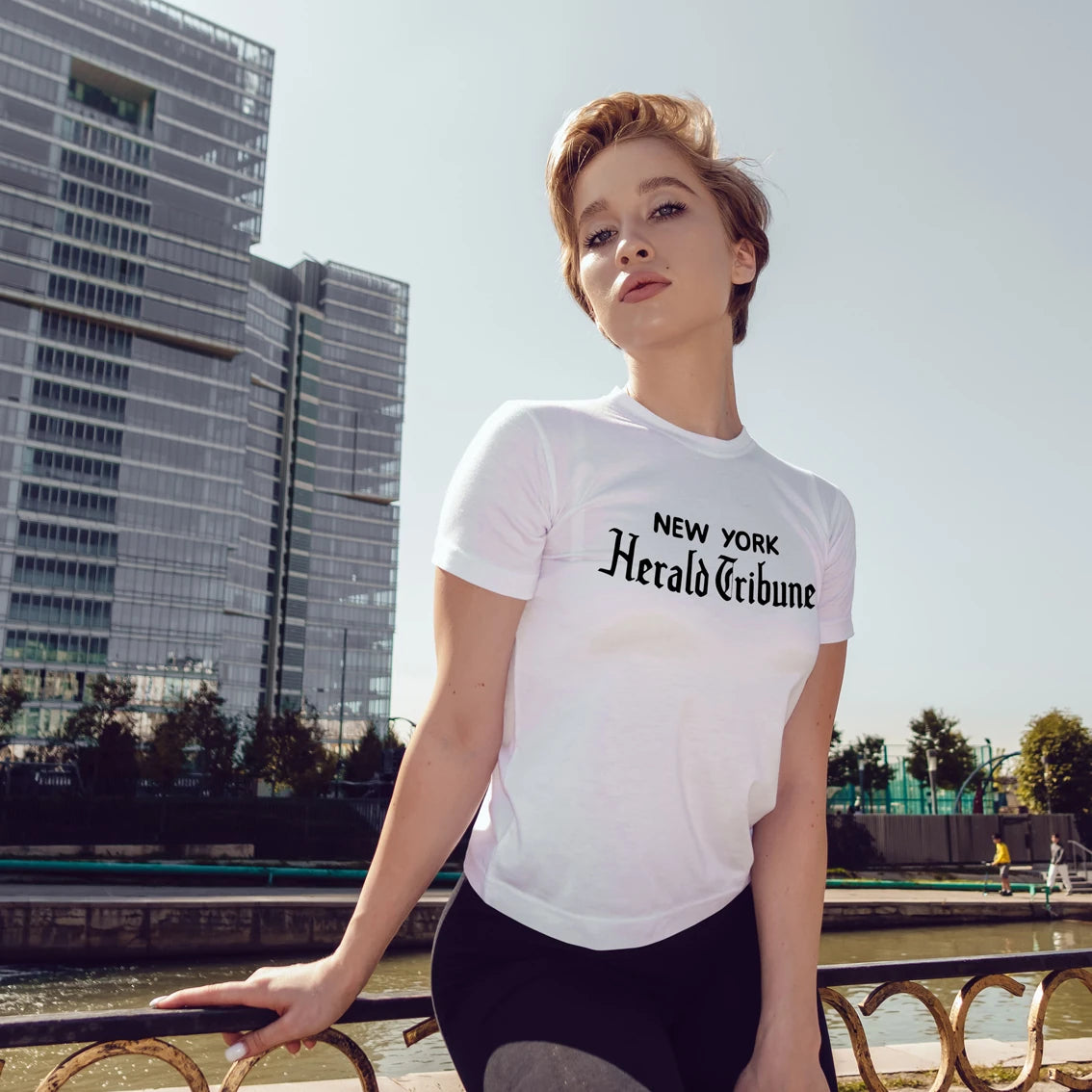 Woman wearing a white t-shirt with 'New York Herald Tribune' text, standing outdoors with modern buildings in the background.
