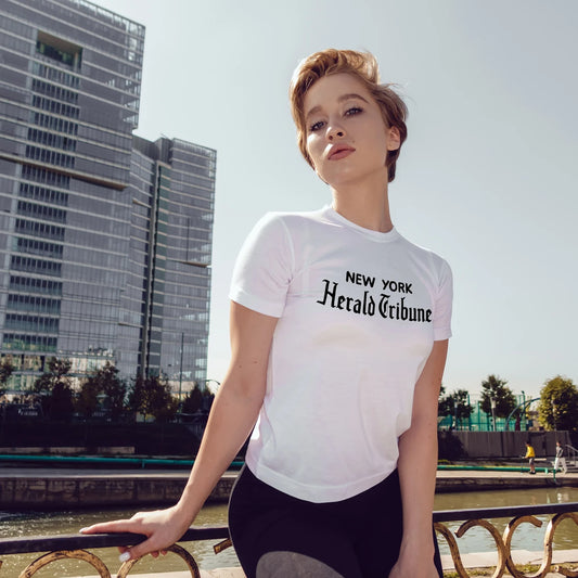 Woman wearing a white t-shirt with 'New York Herald Tribune' text, standing outdoors with modern buildings in the background.