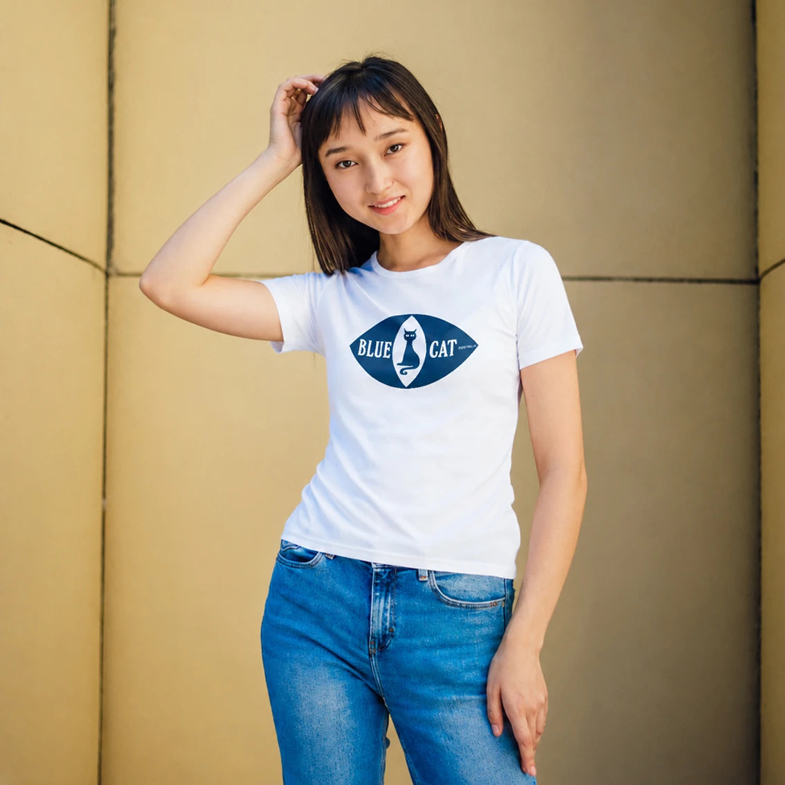 A woman wearing a white short-sleeve t-shirt with a blue 'Blue Cat' cat logo on the chest, paired with blue jeans, standing against a beige paneled wall.