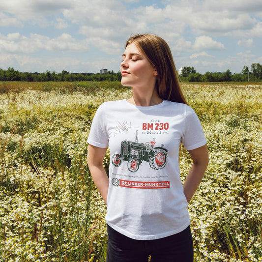 A woman stands in a field of flowers wearing a white t-shirt featuring a vintage Blinder-Munktell BM 230 tractor design. The background shows a scenic outdoor landscape with wildflowers, trees, and a cloudy sky.