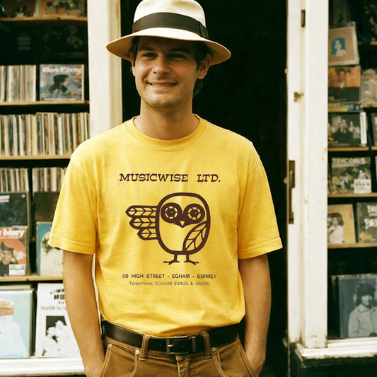 Man wearing a yellow t-shirt with 'Musicwise Ltd.' text and owl logo in a record store.