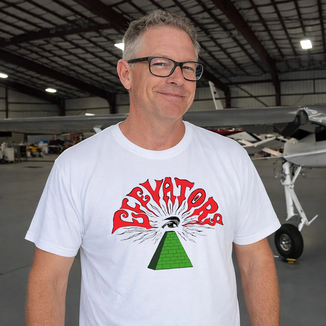 Man in glasses wearing a white t-shirt with a graphic design and 'Elevators' text in an airplane hangar