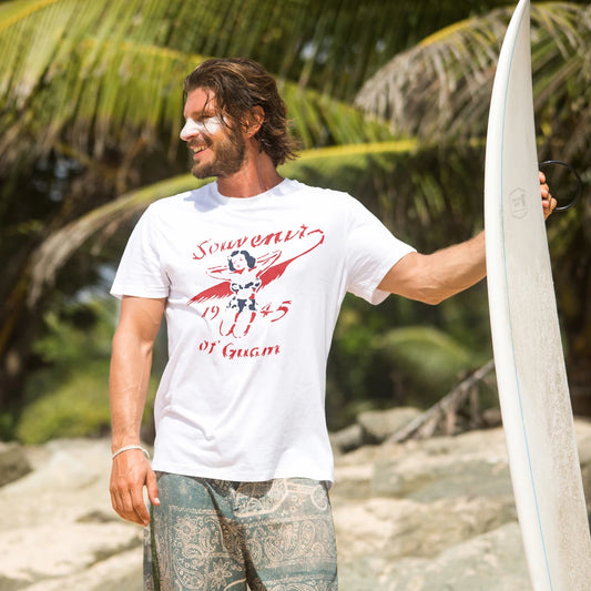 Man holding a surfboard and wearing t-shirt with 'Souvenir Of Guam -1945' text with a tropical background