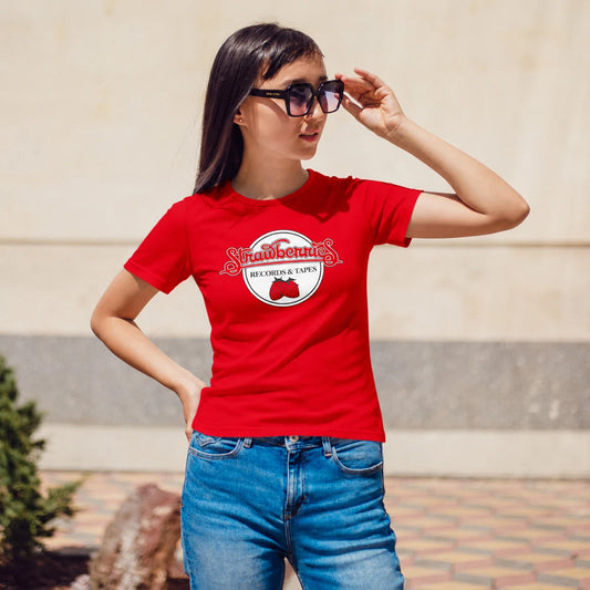 A woman wearing a red graphic t-shirt with a strawberries logo and blue jeans, standing outdoors against a beige stone wall.