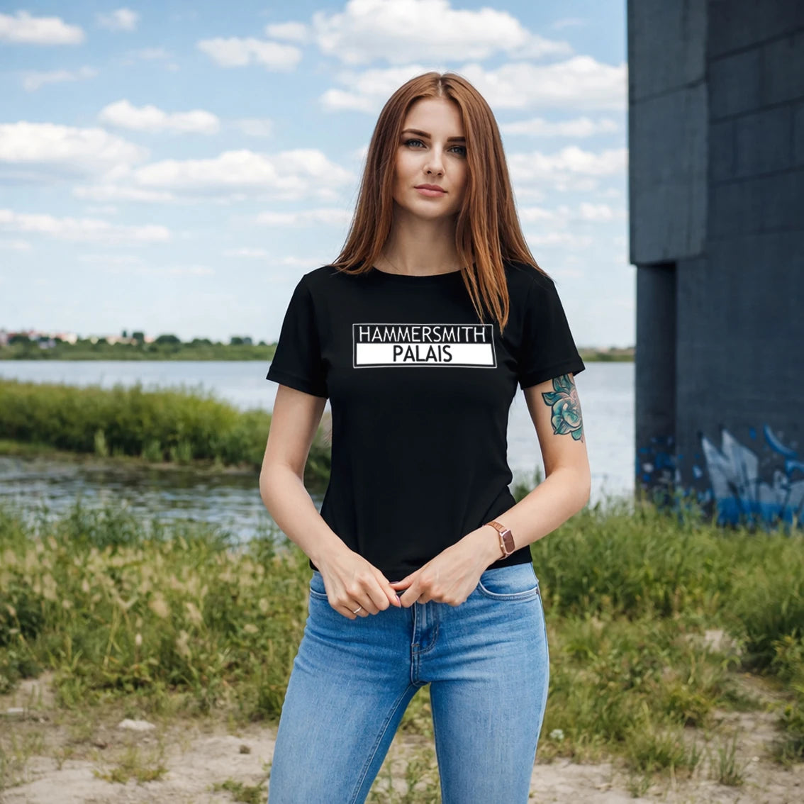 Woman wearing a black t-shirt with 'HAMMERSMITH PALAIS' text, standing outdoors by a body of water.