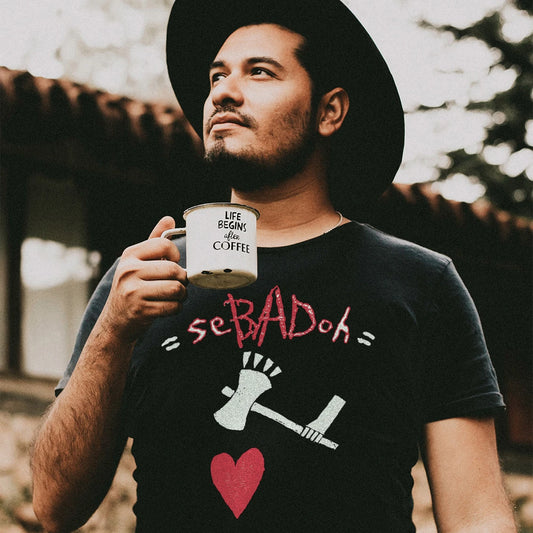 Man holding a mug with 'Life Begins After Coffee' text, wearing a black t-shirt with love heart and axe graphics and 'Sebadoh' text outdoors.