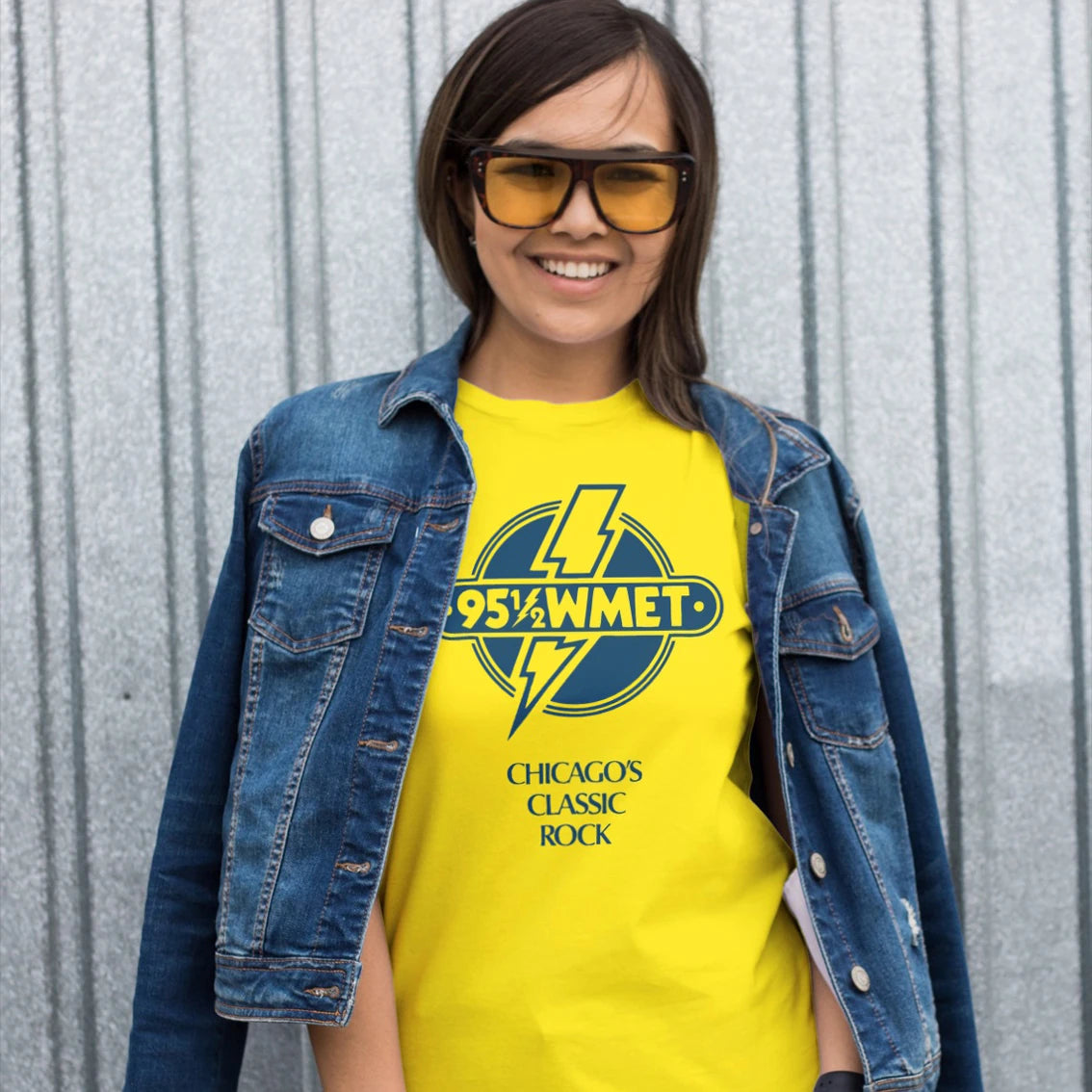 Woman wearing a yellow t-shirt with a radio station logo and '95.5 WMET -CHICAGOS CLASSIC ROCK' text, standing against a corrugated metal background.