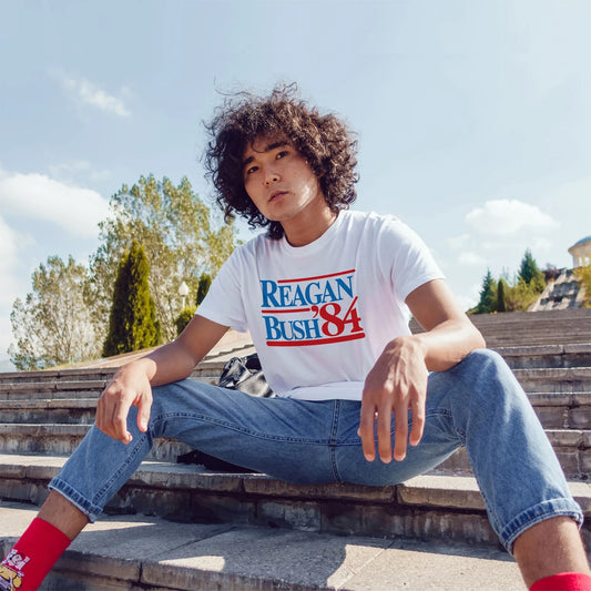 Man wearing a 'Reagan Bush 84' t-shirt sitting outdoors on steps.