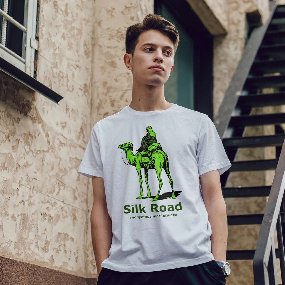 A young man wearing a white t-shirt with a green graphic of a camel & passenger and 'Silk Road' text, standing outdoors.
