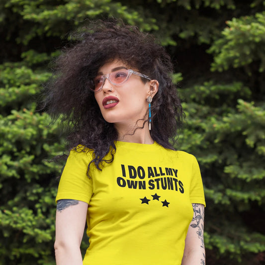 Woman wearing a yellow t-shirt with 'I DO ALL MY OWN STUNTS' text, and three stars design standing against a green foliage background.