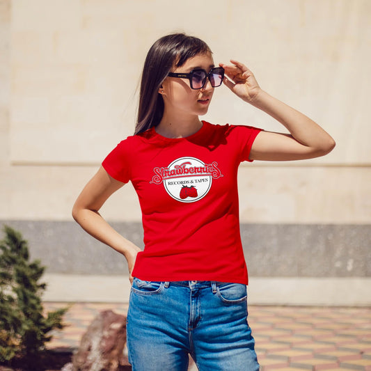A woman in sunglasses is wearing a red t-shirt with a red strawberry on a white circle shape background, saying 'Strawberries Records & Tapes ' standing outdoors.