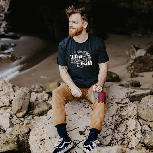 A man is sitting on rocks by a beach wearing a black t-shirt with 'The Fall' logo.