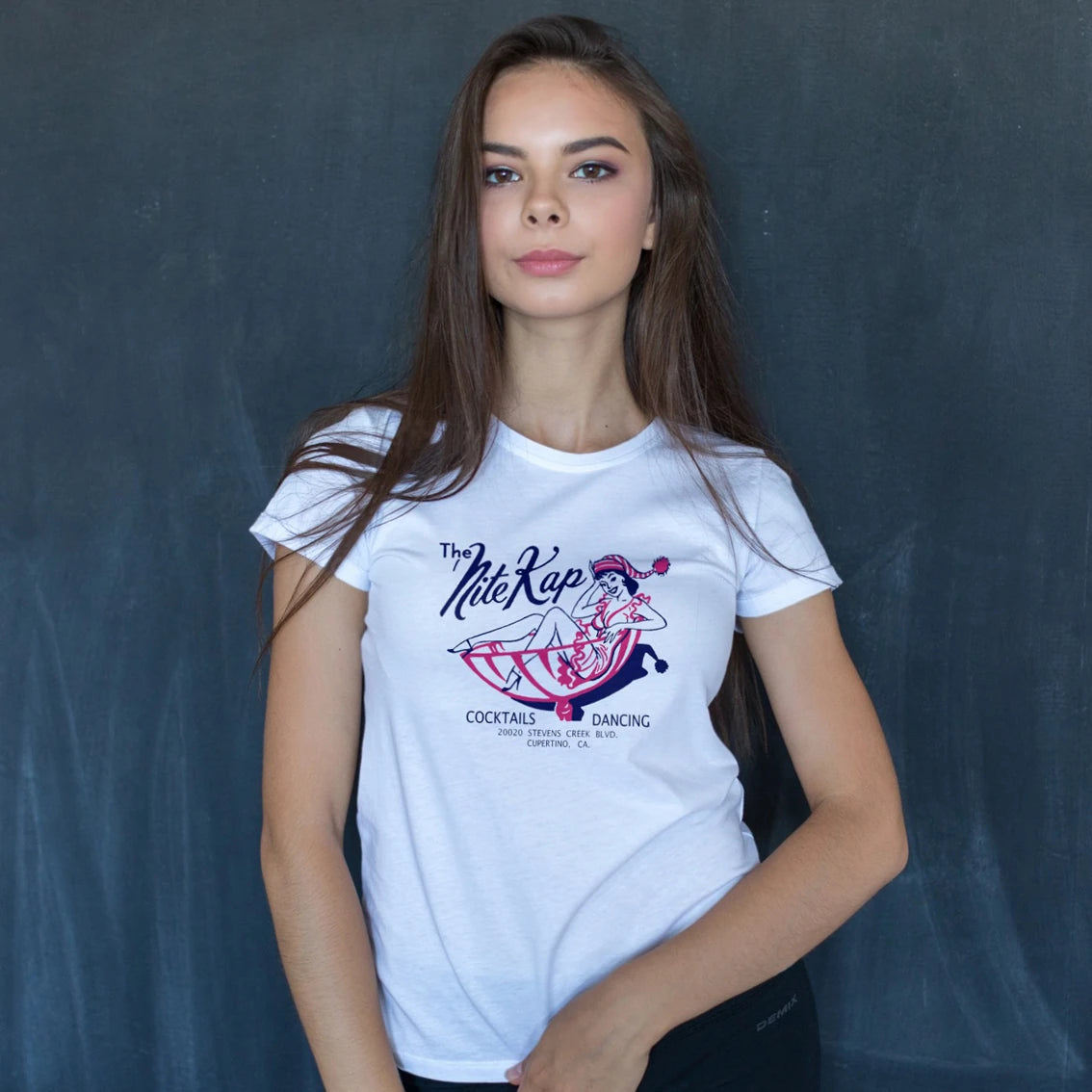 A woman is wearing a white t-shirt with a graphic design text saying 'The Nite Kap - Cocktails, Dancing' and a picture of woman partying on it against a dark background