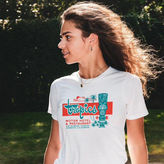 A woman is wearing a white t-shirt with a colorful tropical looking graphic design and text saying 'Tropics -  Motor Hotel & Restaurant' standing outdoors.