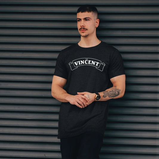 A man with a mustache is wearing a black t-shirt with 'VINCENT' logo printed on it, standing against a metallic shutter background.