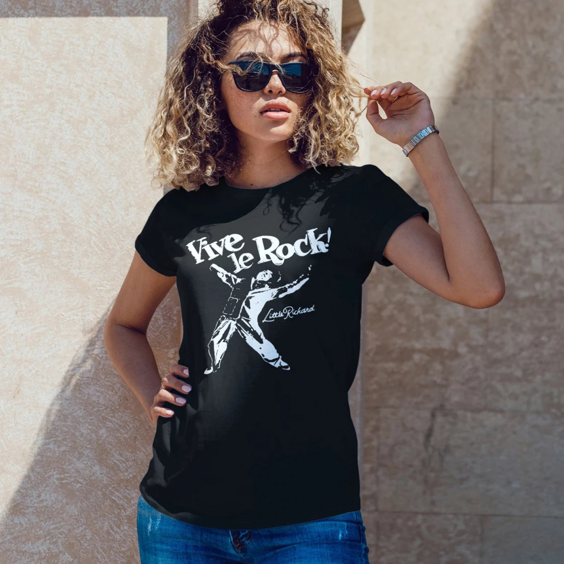 A woman in sunglasses is wearing a black t-shirt with 'Live Le Rock' text and picture of the man, standing against a stone wall.