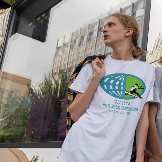 A person is wearing a white t-shirt with a graphic design of 'ISF/USSA - WORLD SURFING CHAMPIONSHIPS', standing in an urban setting