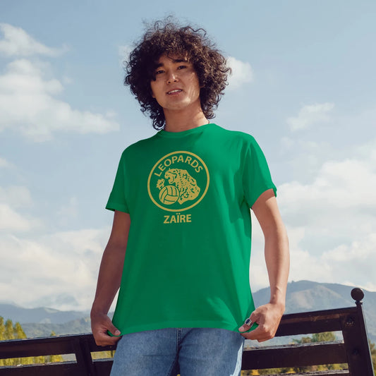 A man is wearing a green t-shirt with a logo of leopard & ball, and 'Leopards, ZAIRE' text, standing outdoors with a scenic background.